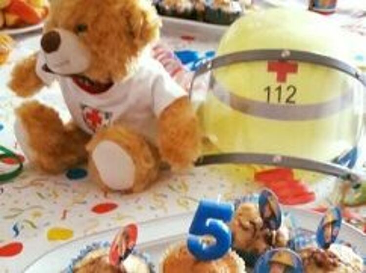 DRK-Kindergeburtstag Muffins mit einer 5 sind auf einer Geburtstagsdecke angerichtet, ein DRK-Helm und ein Trösteteddy daneben